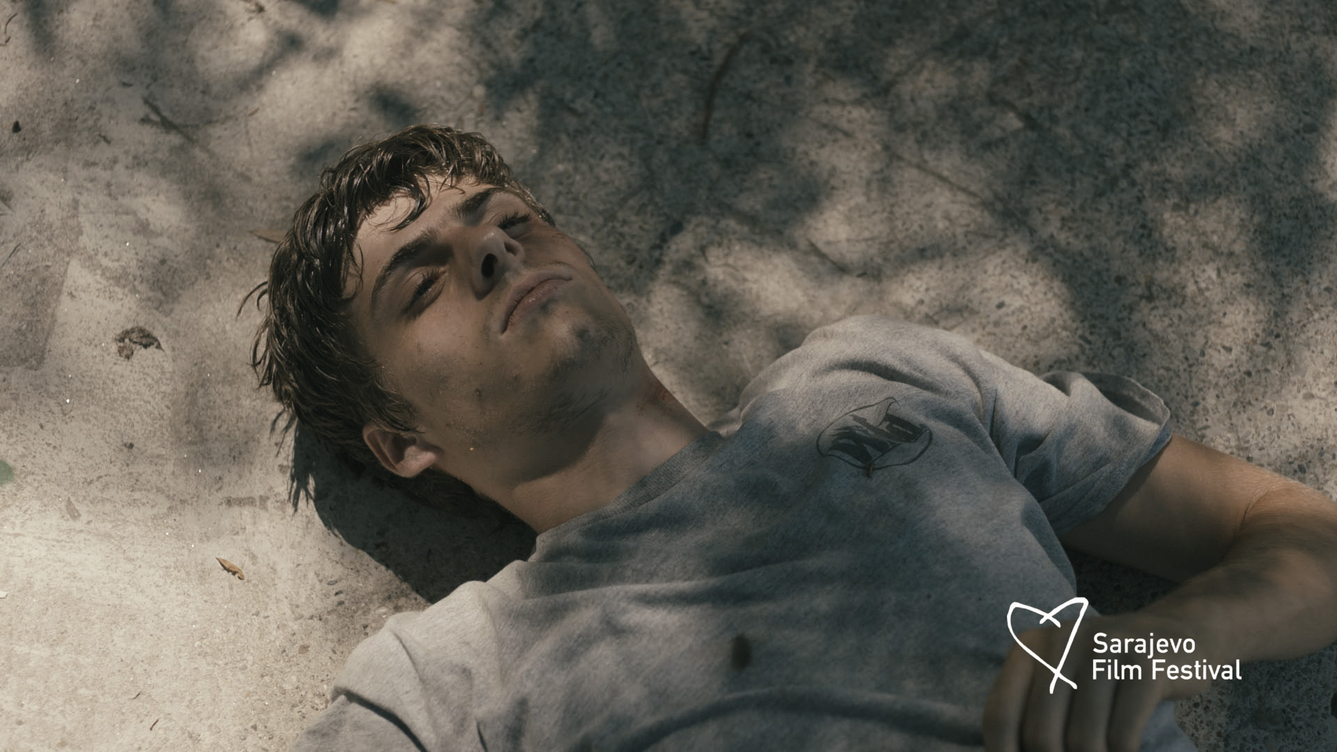 Young man in a gray t-shirt lying on a rough concrete surface with shadows overhead.