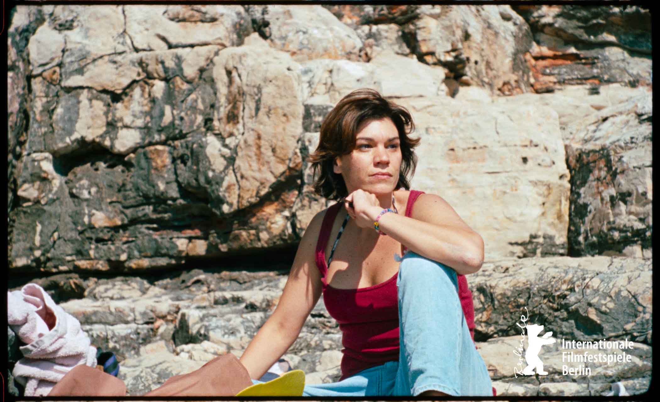 Woman wearing a red tank top and blue jeans sitting on rocks, looking contemplative outdoors.