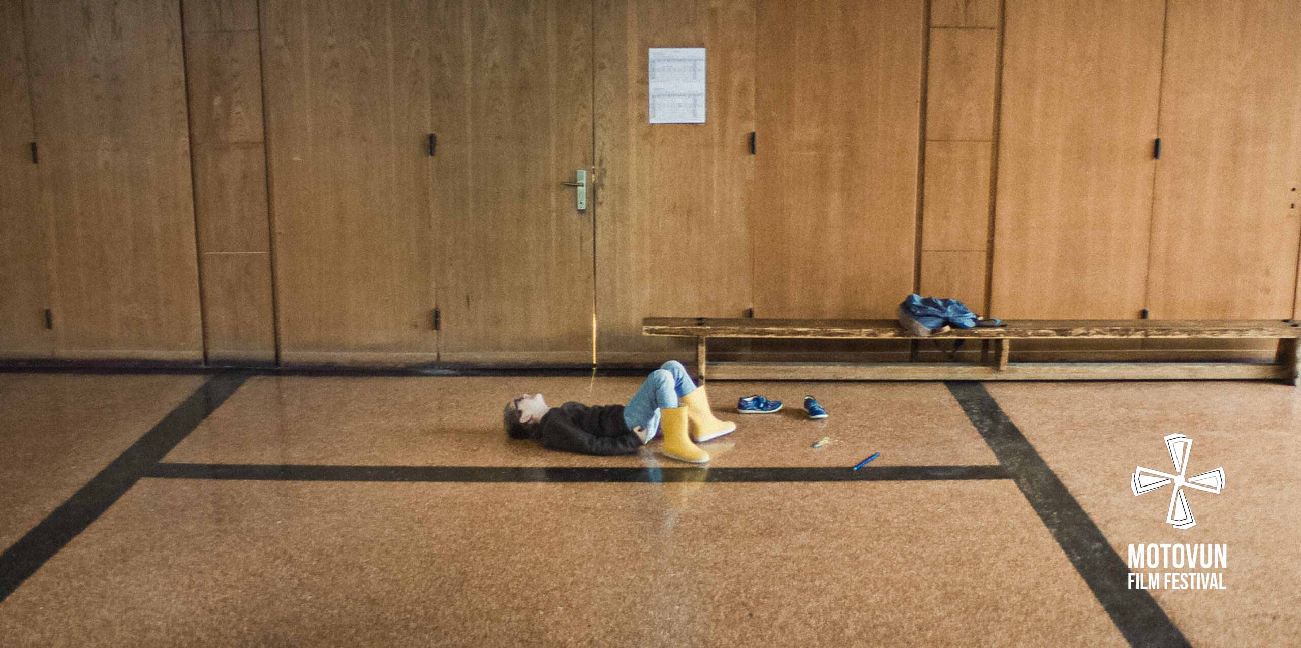 Child in yellow boots lying on a gym floor near wooden benches and a door with papers taped on it.