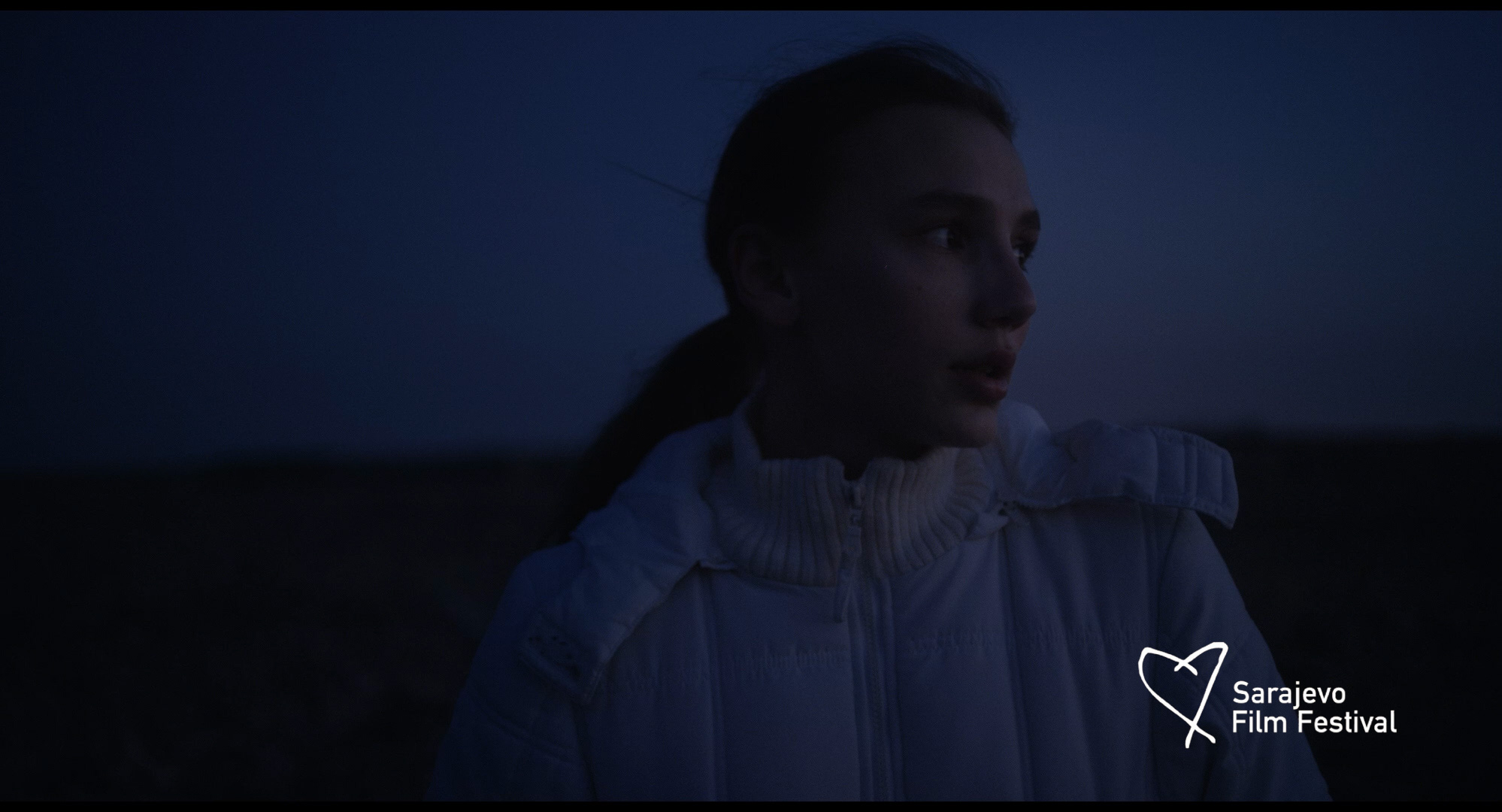 Young woman in white jacket looking to the side against a dark, dusk sky background.