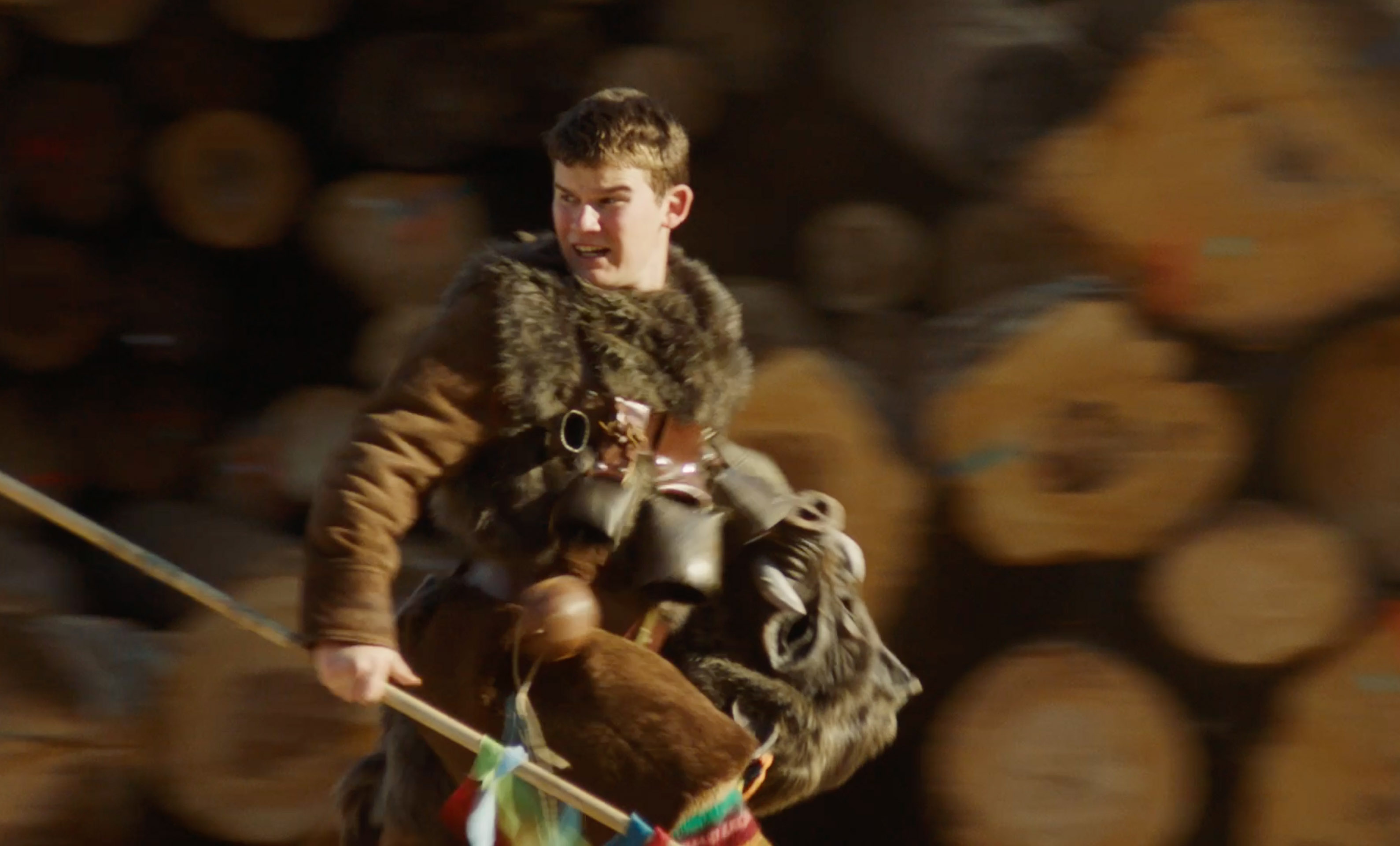 Young man in fur vest holding a stick and a boar's head mask with stacked logs background.
