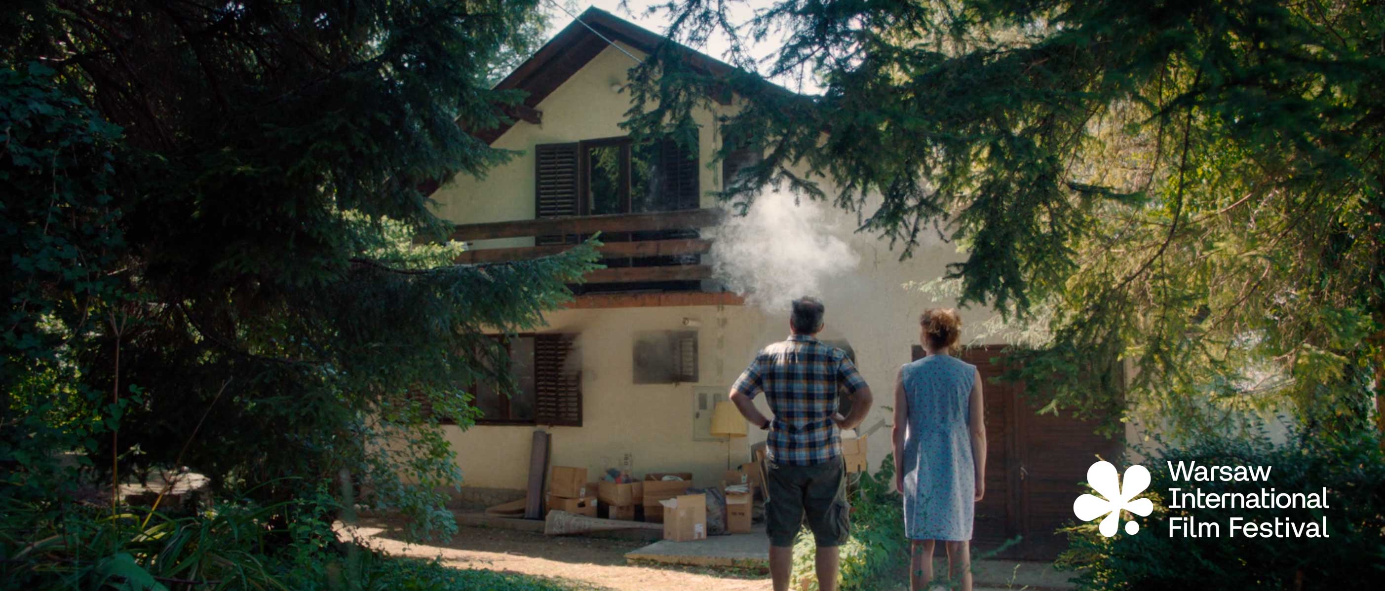 Man and woman stand facing a house with smoke coming from a window among trees and boxes outside.