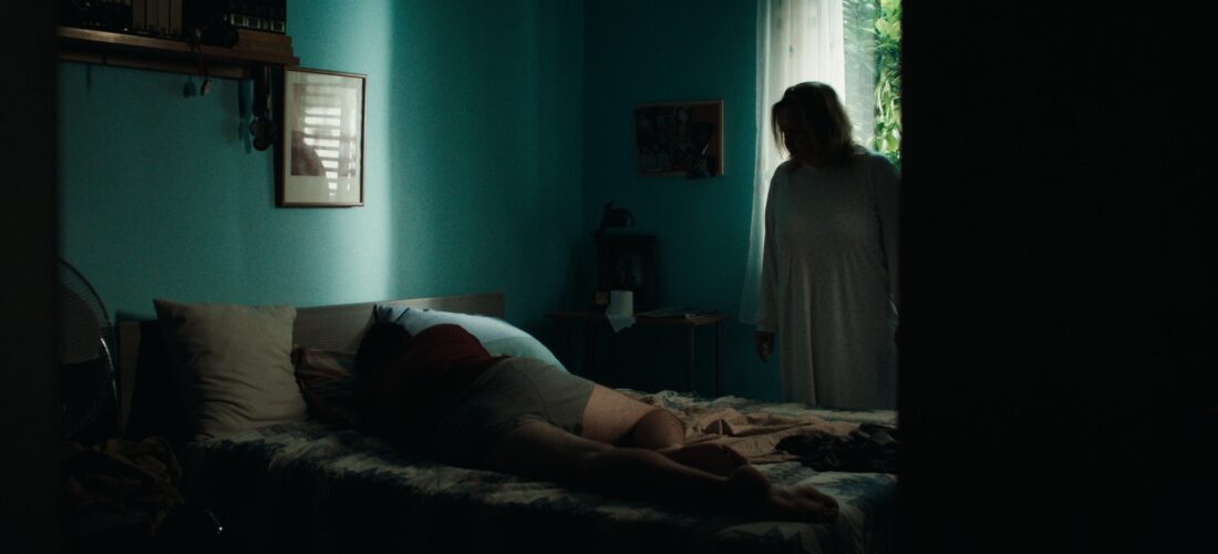 A person lying face down on a bed with another standing near a window in a dimly lit room.