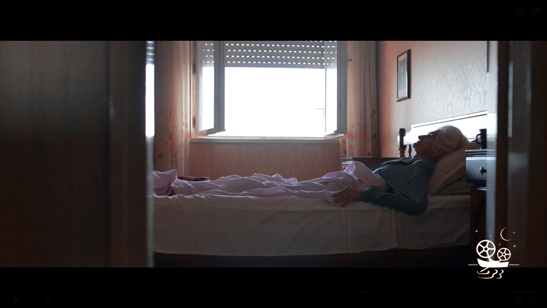 Elderly woman lying in bed by window in softly lit room with floral curtains and wallpaper.